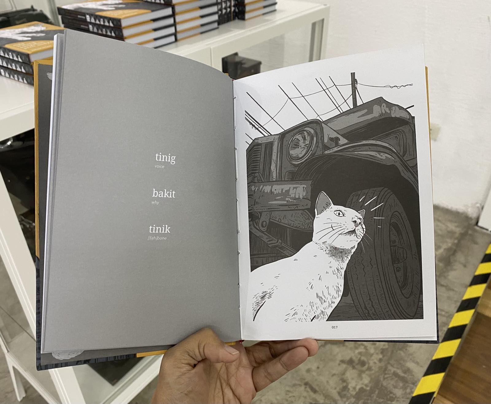 Text on the left: "tinig (voice), bakit (why), tinik ((fish)bone)". On the right is a white cat with a close up of a grey car in the background. We see the cat from a bug's perspective; the cat is looking up, having seen something that caught its attention. Page 017.